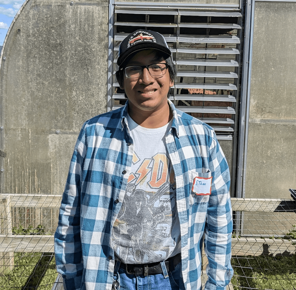 Diversified Vegetable Pre-Apprenticeship graduate Ethan Kramer in a black hat and blue flannel smiling at the camera in front of a greenhouse